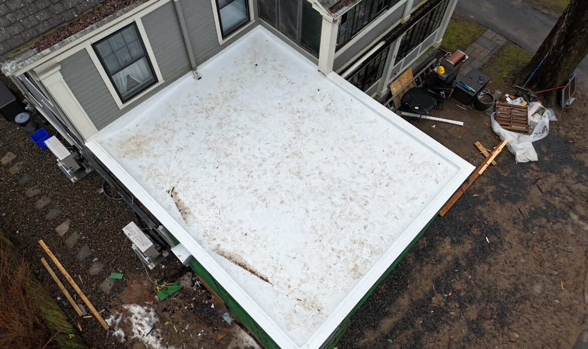 Aerial view of a new white membrane flat roof on a one-story addition attached to a gray-sided house