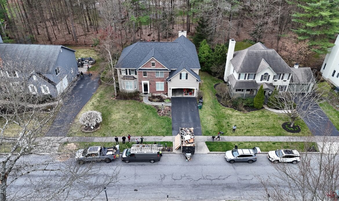 Drone view of a brick-front colonial with a dumpster of torn-off shingles and crew trucks parked on the street