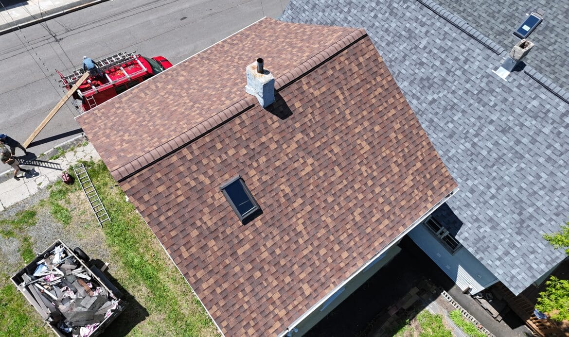 Brown architectural shingle roof with a skylight and chimney, crew loading ladders at the street below