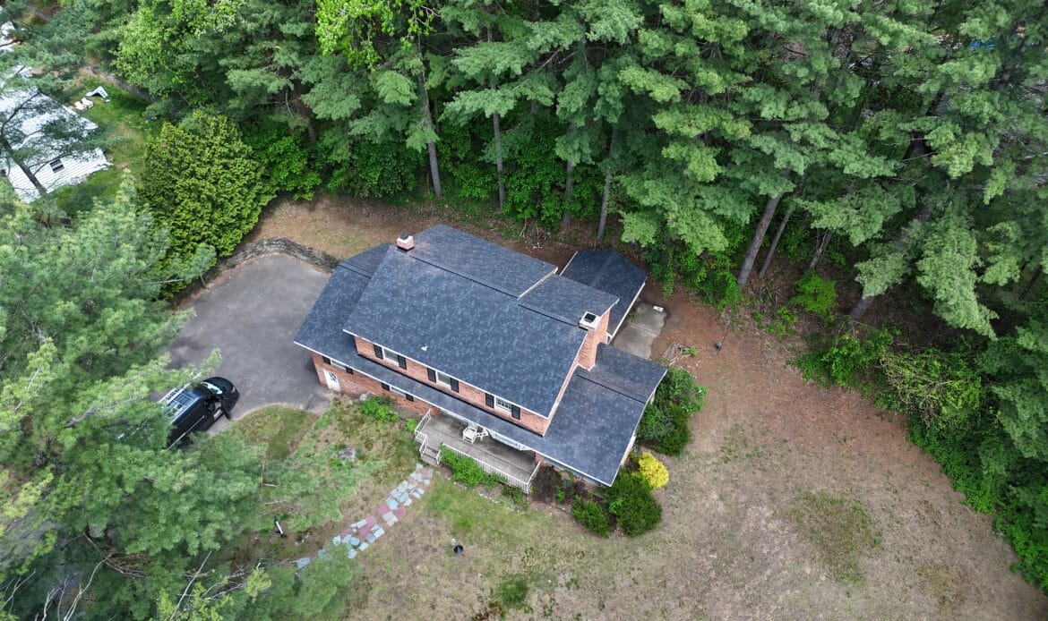 Aerial view of a brick two-story house with a dark shingle roof surrounded by tall pine trees