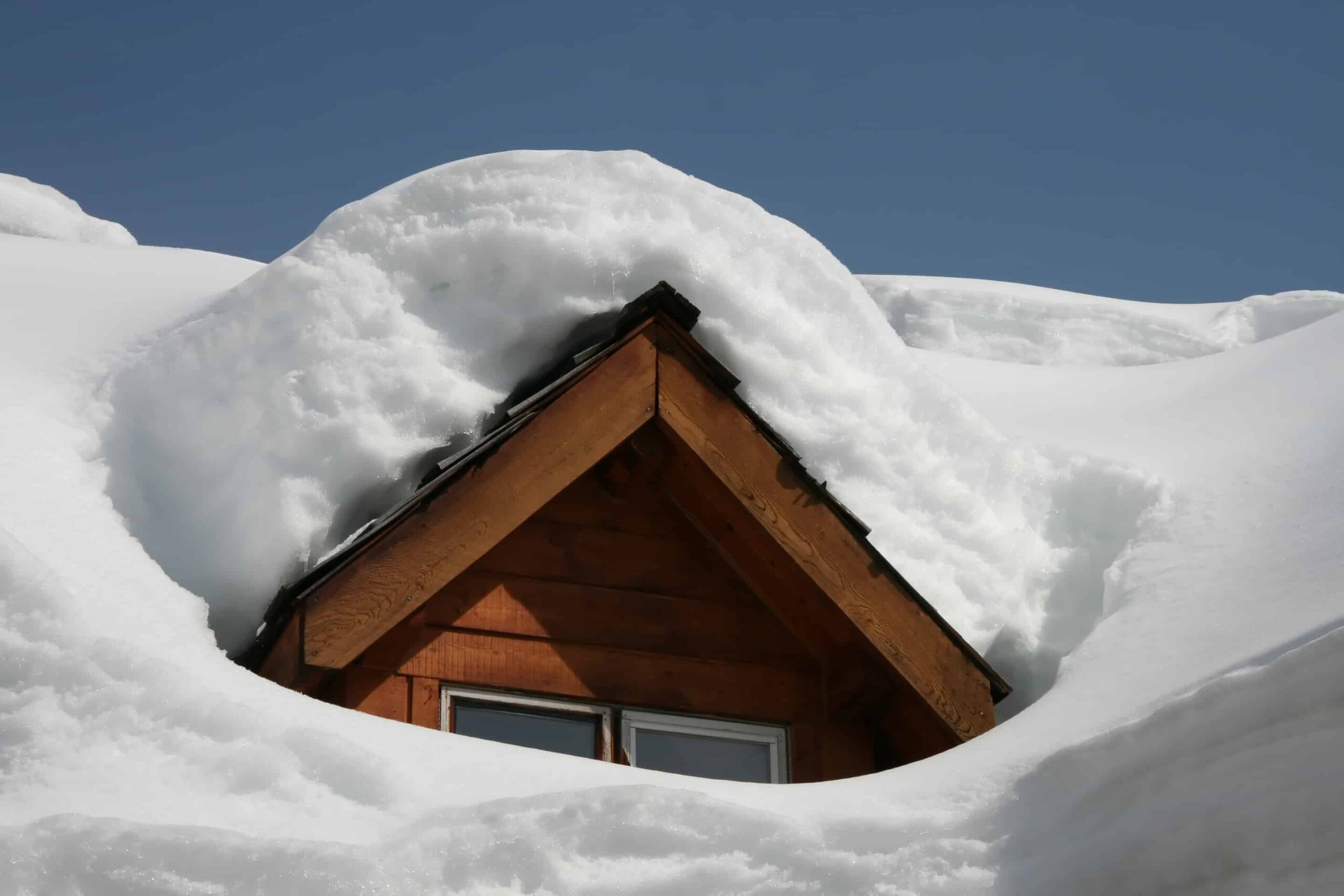 Snow piled over the peak of a roof and dormer on a wood-sided house
