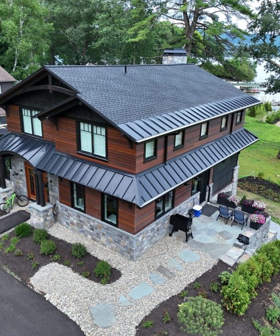 Drone view of a two-story house with dark architectural shingles on the main roof and black standing seam metal on the lower roofs